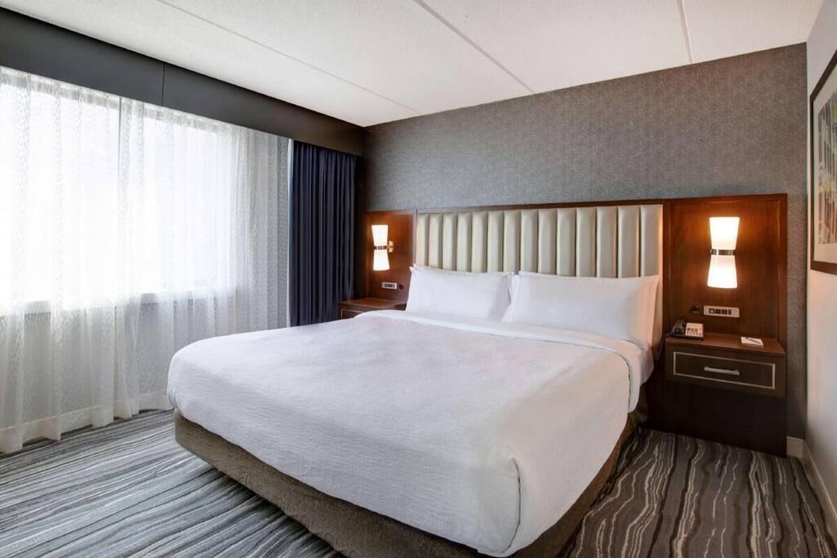 A hotel room with a large bed, two bedside tables with lamps, a headboard, and a window with curtains.
