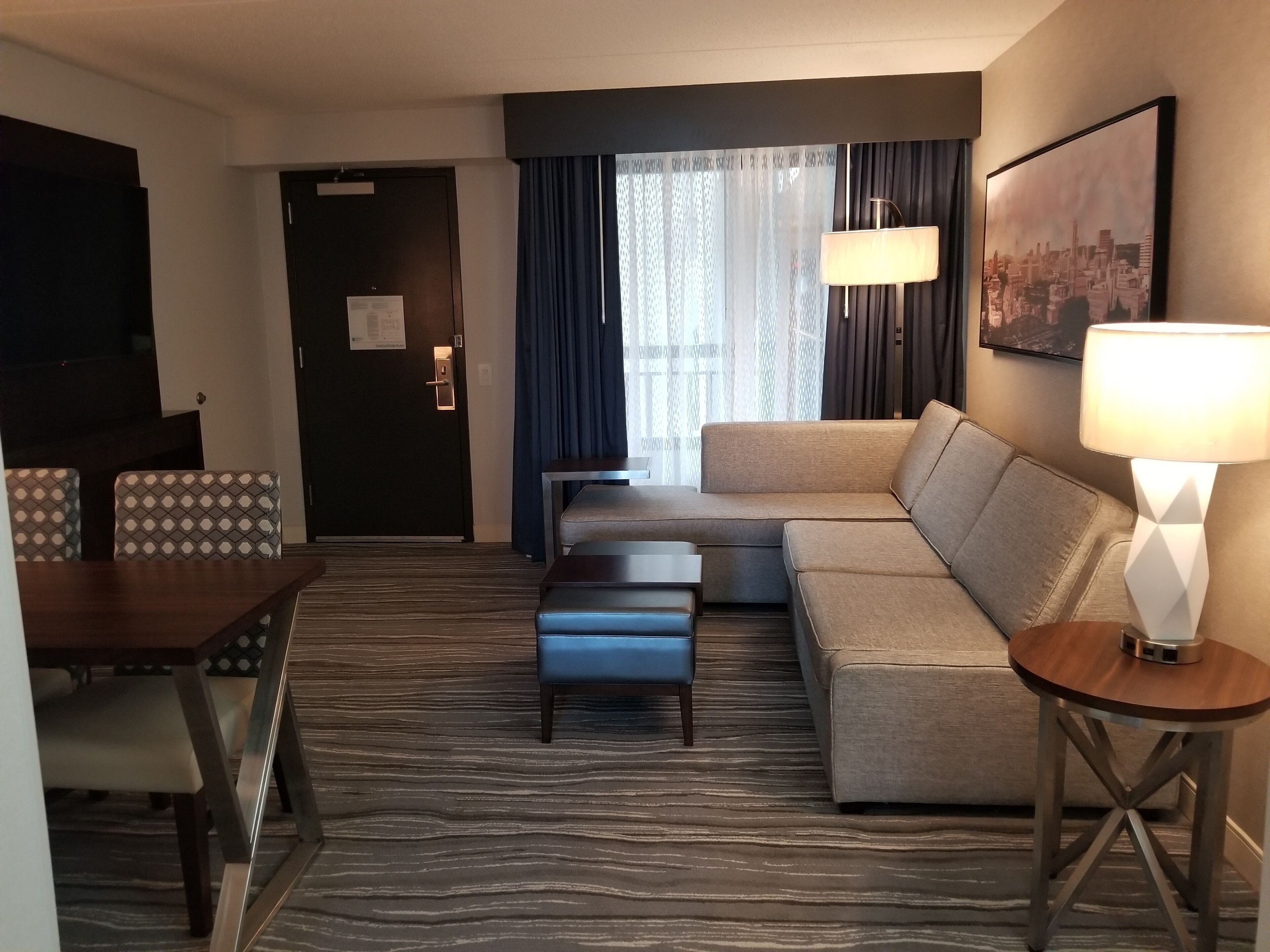 A hotel room with a sofa, a dining table with chairs, a small coffee table, a lamp, and a cityscape painting on the wall.