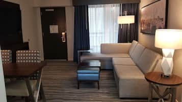 A hotel room with a sofa, a dining table with chairs, a small coffee table, a lamp, and a cityscape painting on the wall.