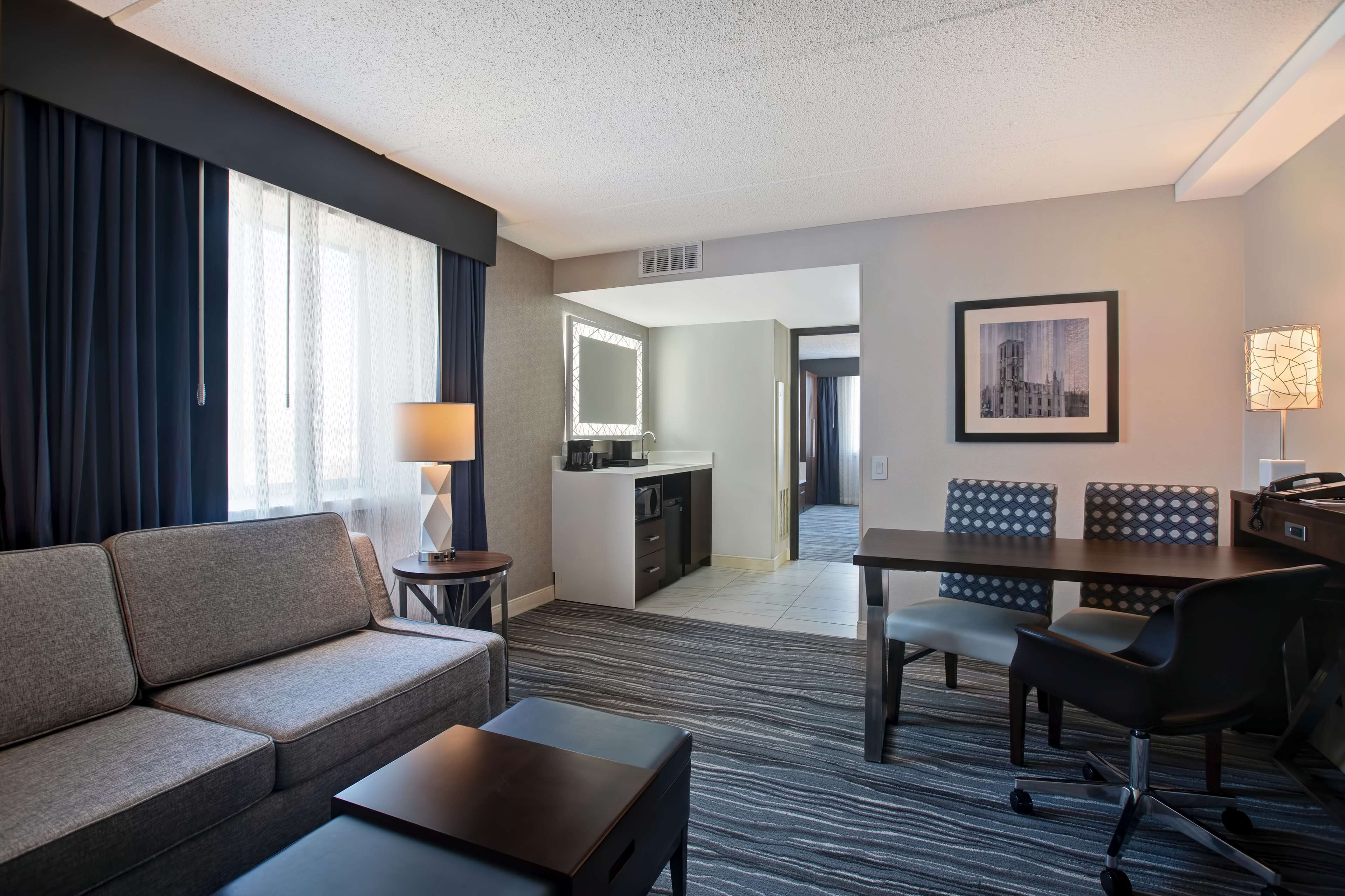 A hotel room with a sofa, a small table, a desk with chairs, a kitchenette, and a dining area.