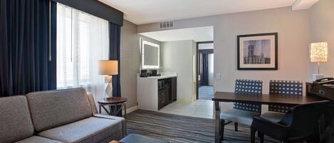 A hotel room with a sofa, a small table, a desk with chairs, a kitchenette, and a dining area.