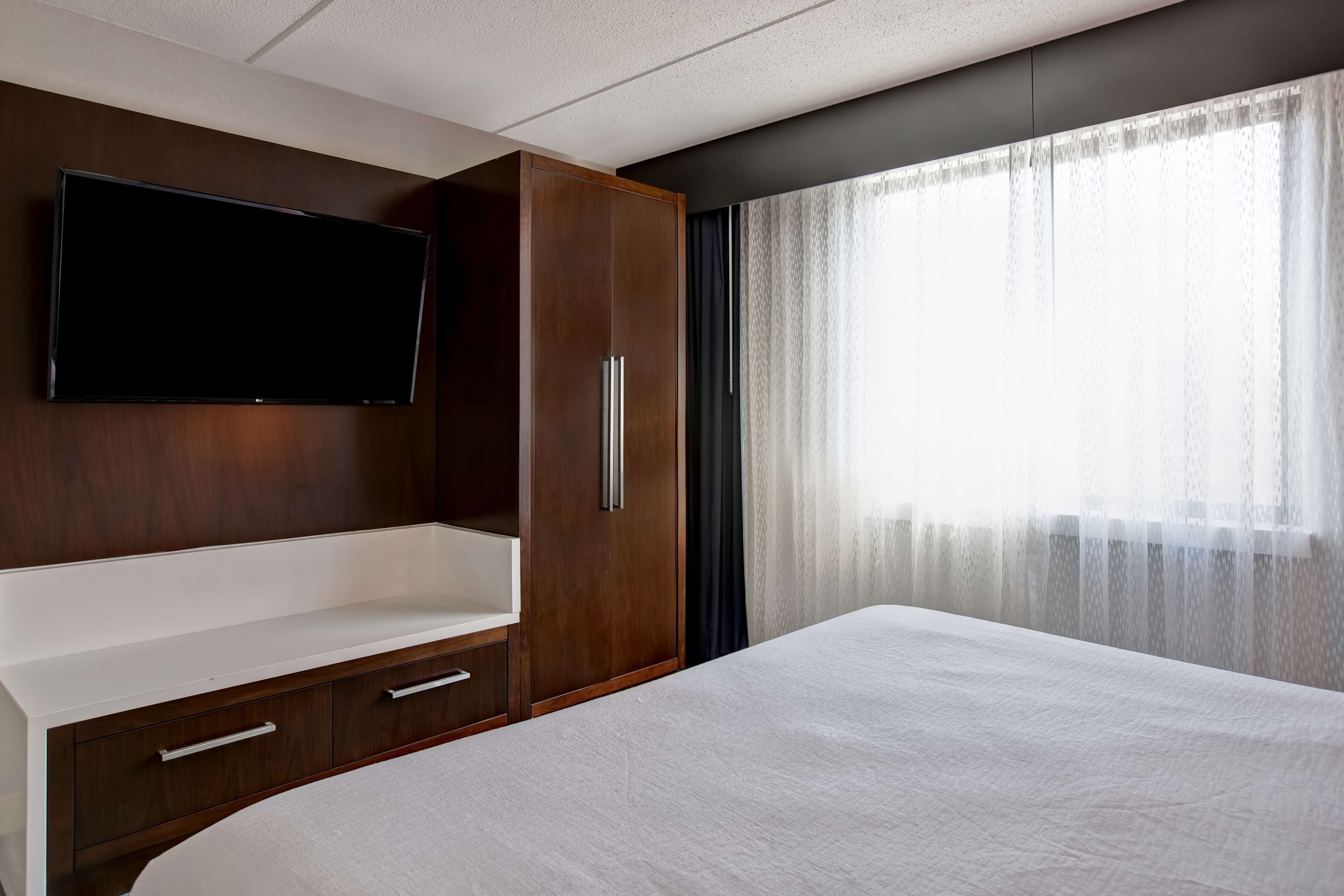 A modern hotel room with a large bed, a built-in TV, a wooden wardrobe, and a large window with sheer curtains.