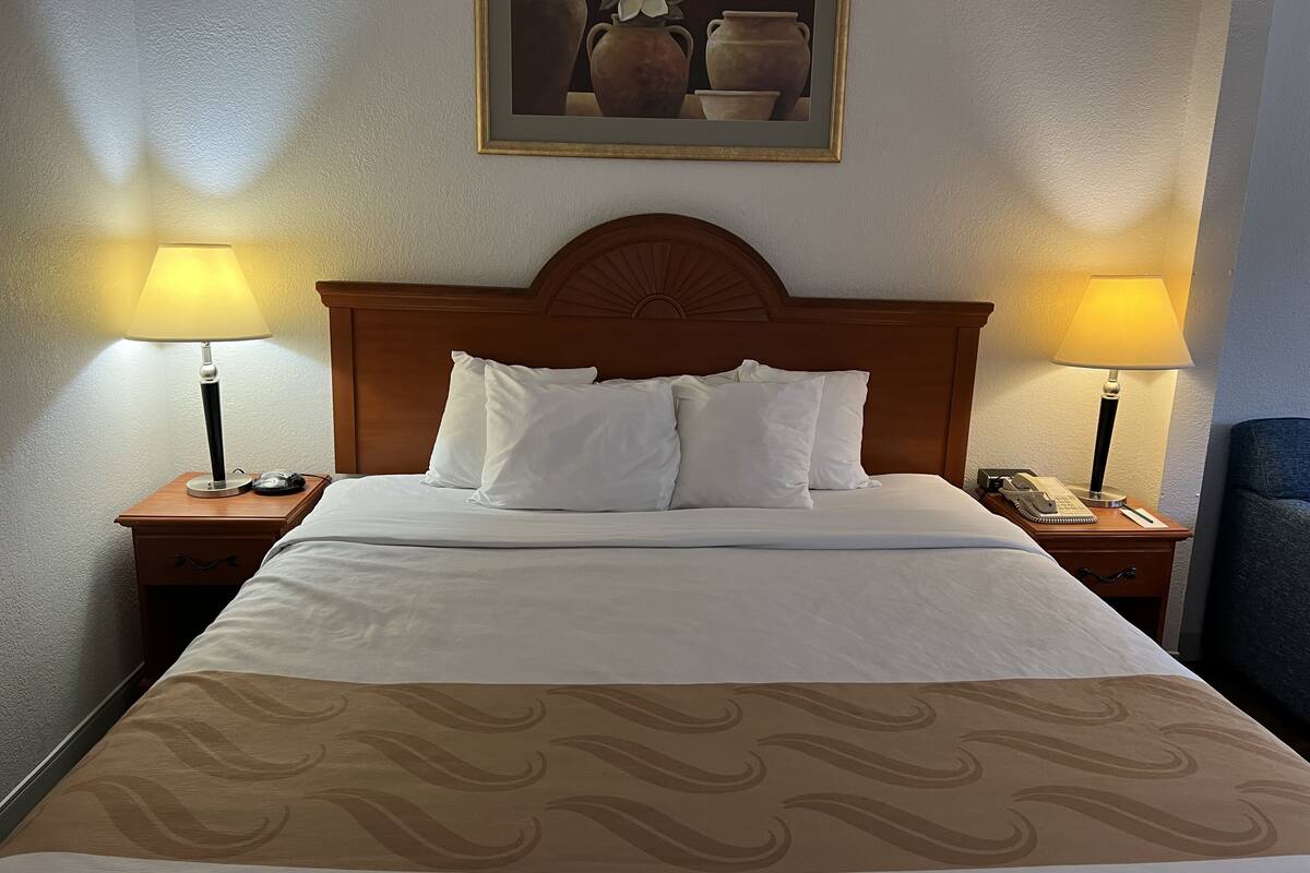 Ein ordentlich bezogenes Bett mit weißen Leinen und einer gemusterten Tagesdecke, flankiert von zwei Nachttischen mit Lampen, einem Schreibtischchen mit Telefon und einem Bild über dem Bett.