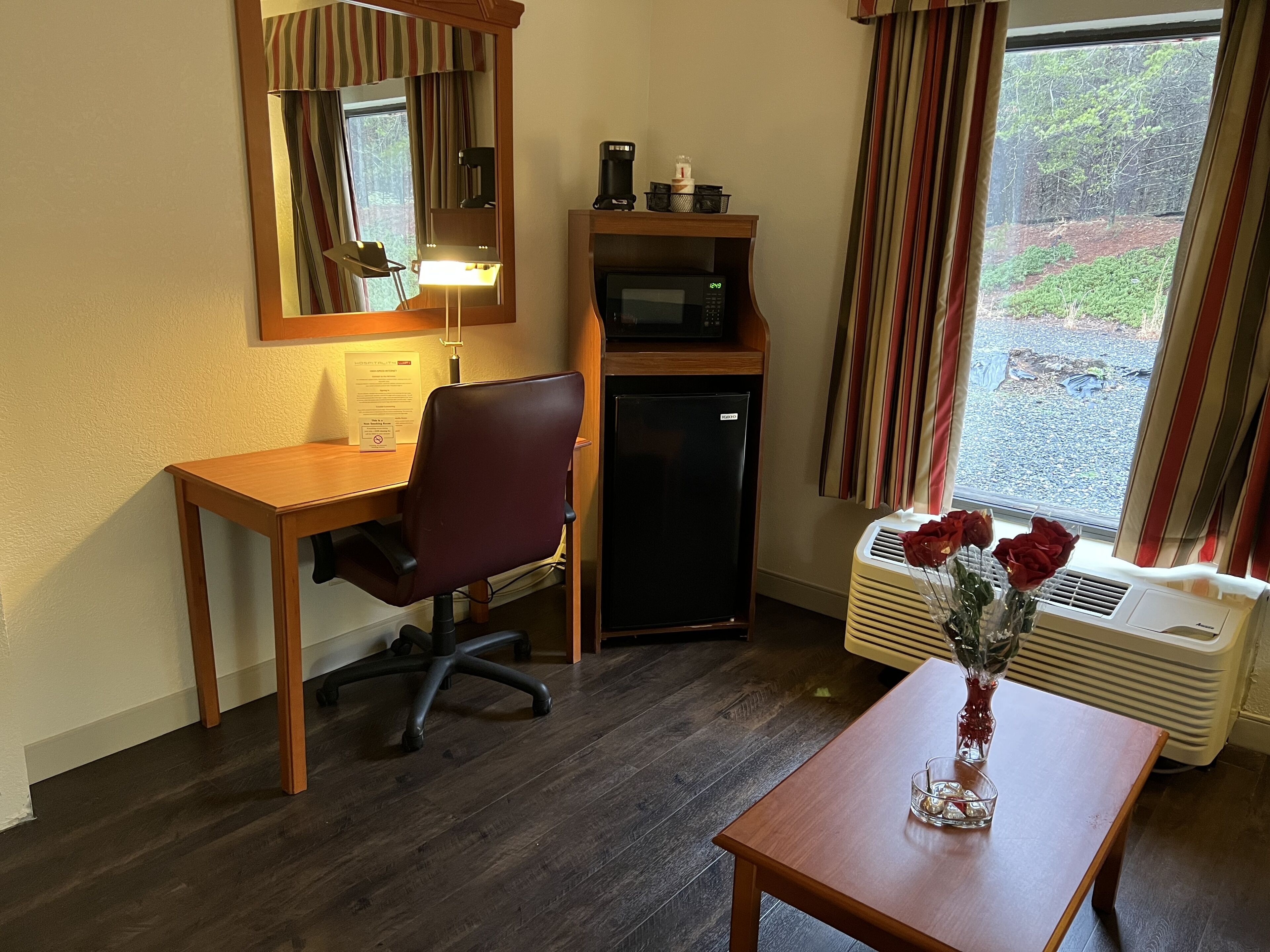 Una habitación compacta con escritorio de madera, una silla, refrigerador pequeño, microondas y ventana con cortinas.