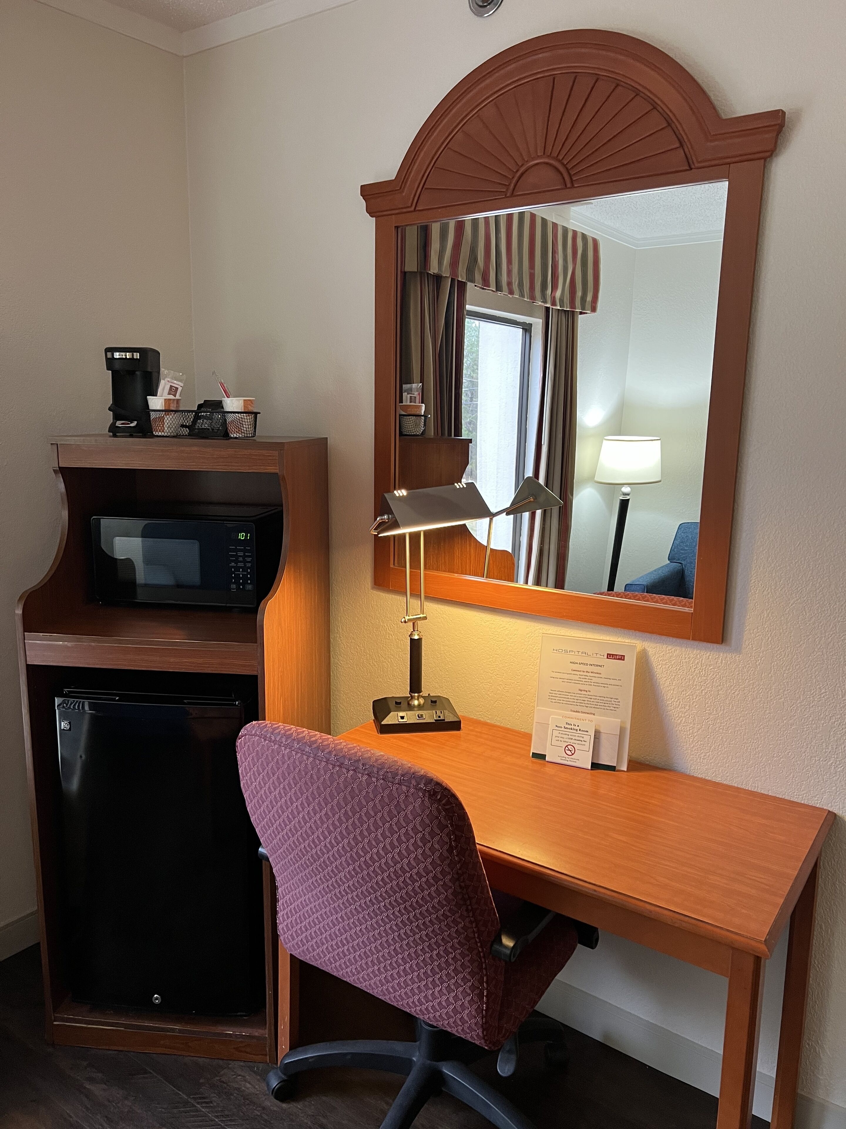 A hotel room with a wooden desk, a purple chair, a microwave, a coffee maker, a lamp, and a mirror.
