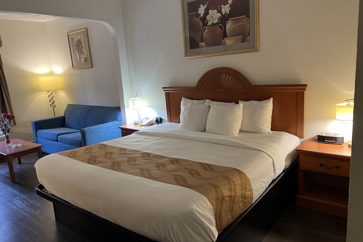 Ein Hotelzimmer mit einem großen Bett, einem blauen Sessel, einem Nachttisch aus Holz mit einer Lampe und einem Bild an der Wand.