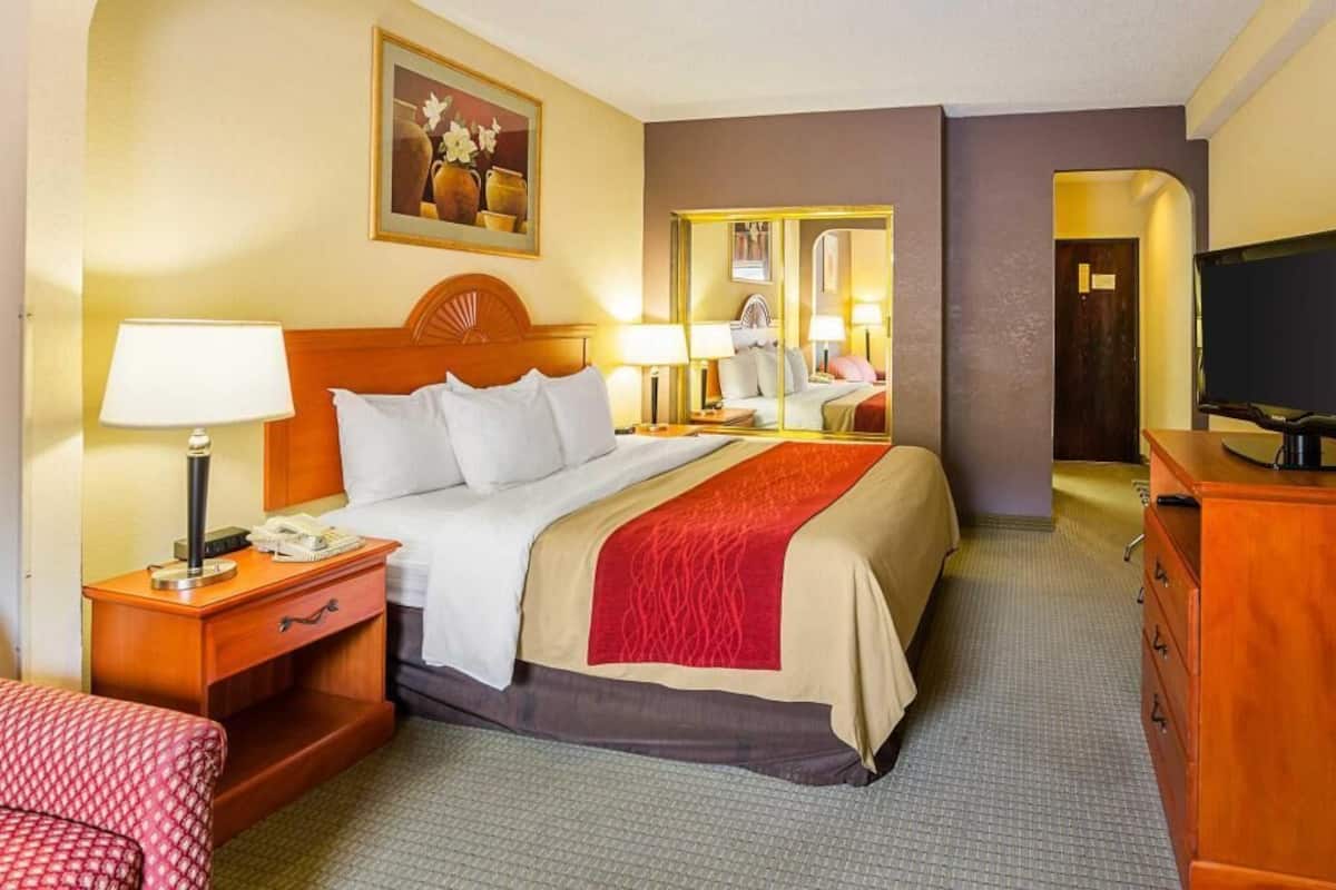 Ein Hotelzimmer mit einem Bett, einem Nachttisch mit Lampe, einem Fernseher, einem Schminktisch und einem Spiegel.