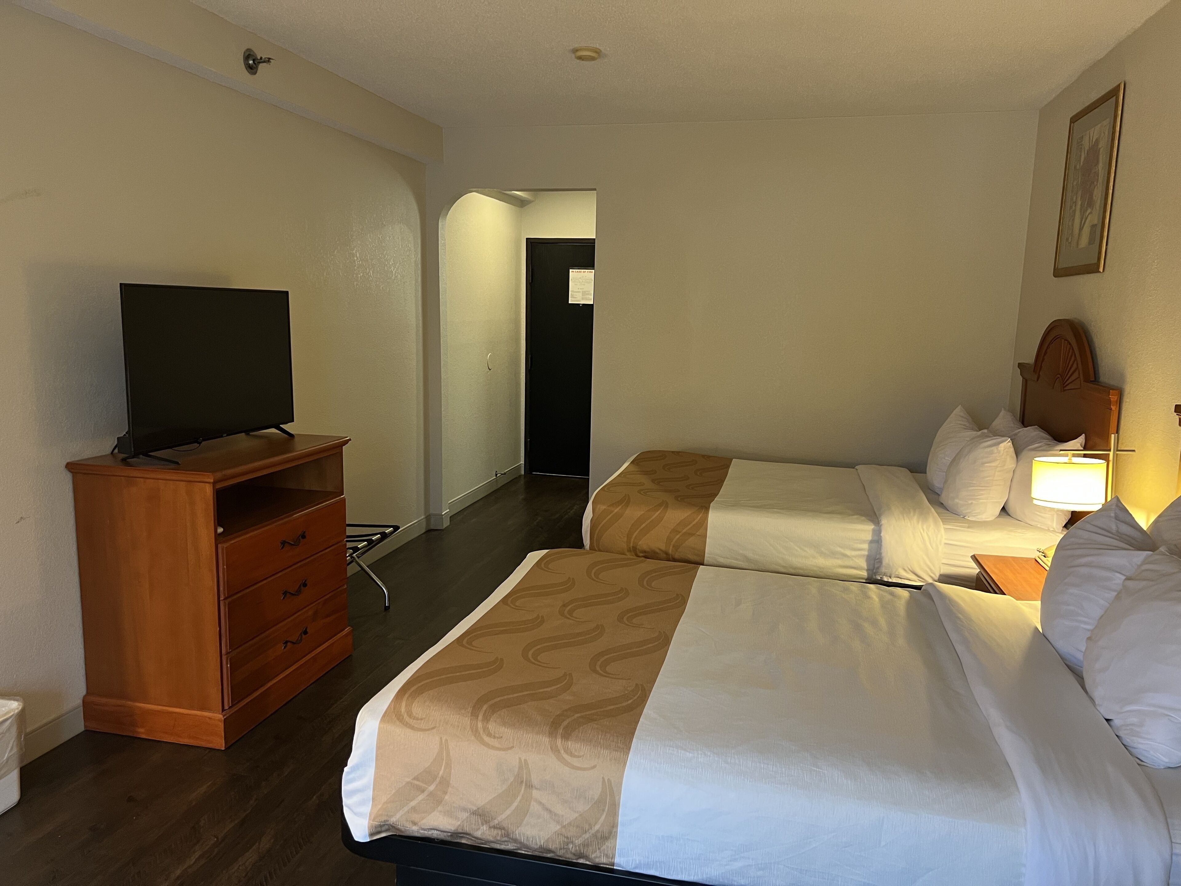 A hotel room with two beds, a TV on a wooden dresser, and a hallway leading to another room.