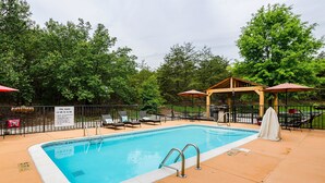 Una piscina con sillones de descanso y sombrillas, rodeada por una valla y árboles.