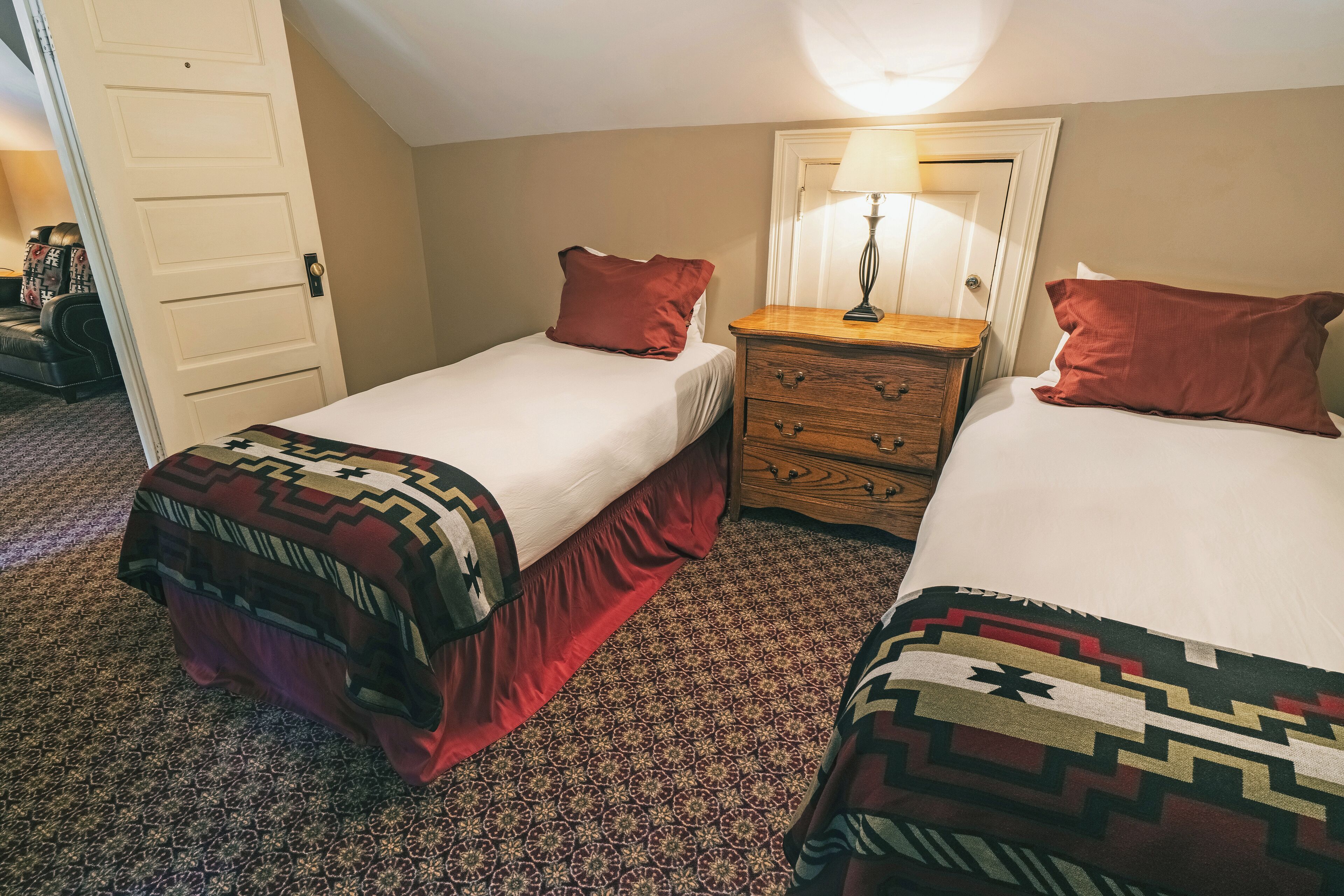 Habitación con dos camas, cada una con un edredón estampado, una mesita de noche de madera con una lámpara y una puerta que da a otra habitación.