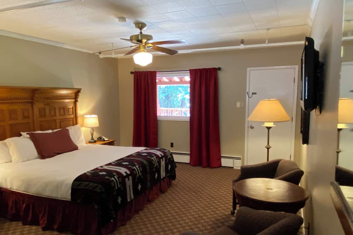 A hotel room with a bed, a ceiling fan, a window with red curtains, a desk with a lamp, and a chair.