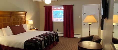 A hotel room with a bed, a ceiling fan, a window with red curtains, a desk with a lamp, and a chair.