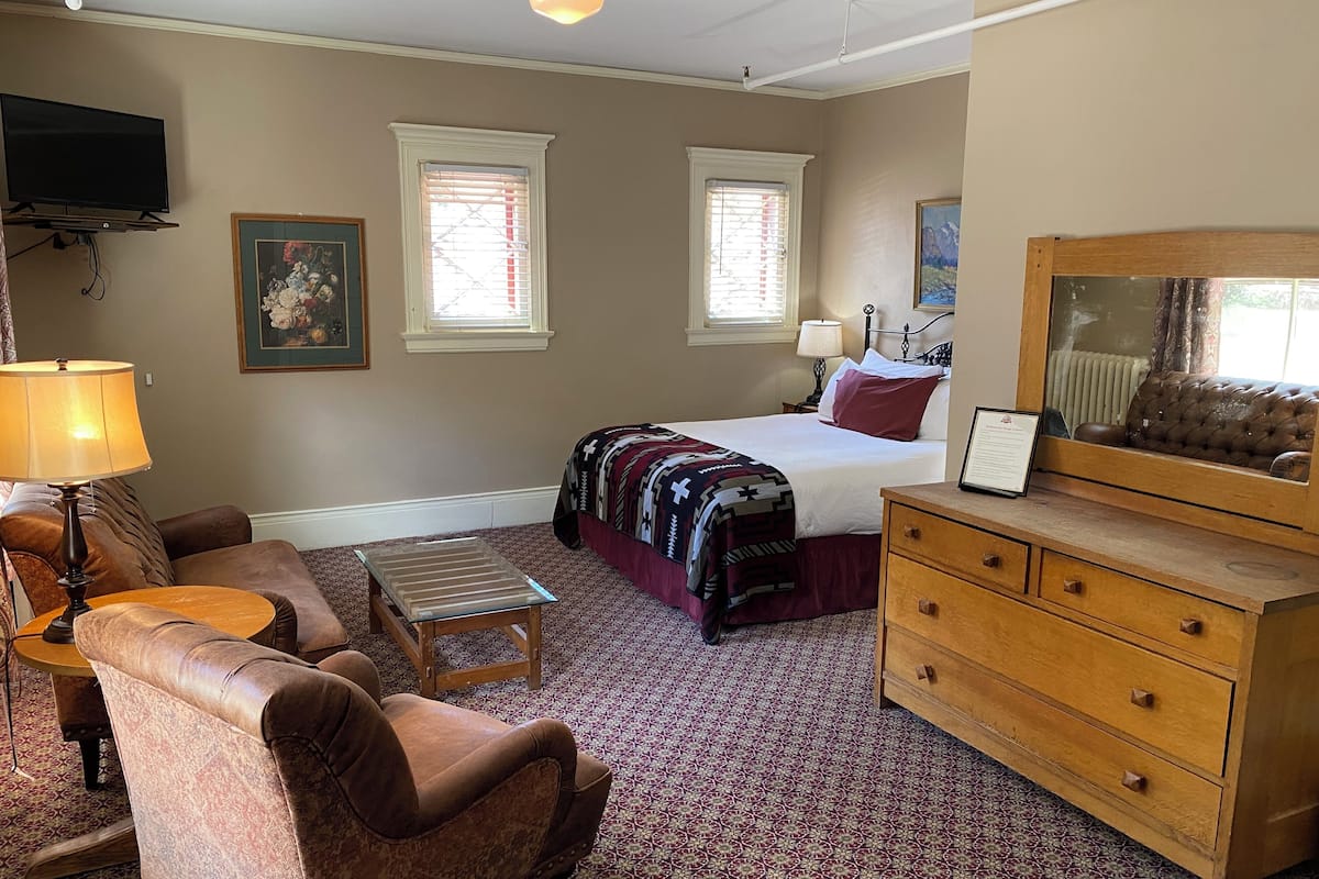 A hotel room with a bed, a wooden dresser, a leather armchair, a small table, a lamp, and a television.