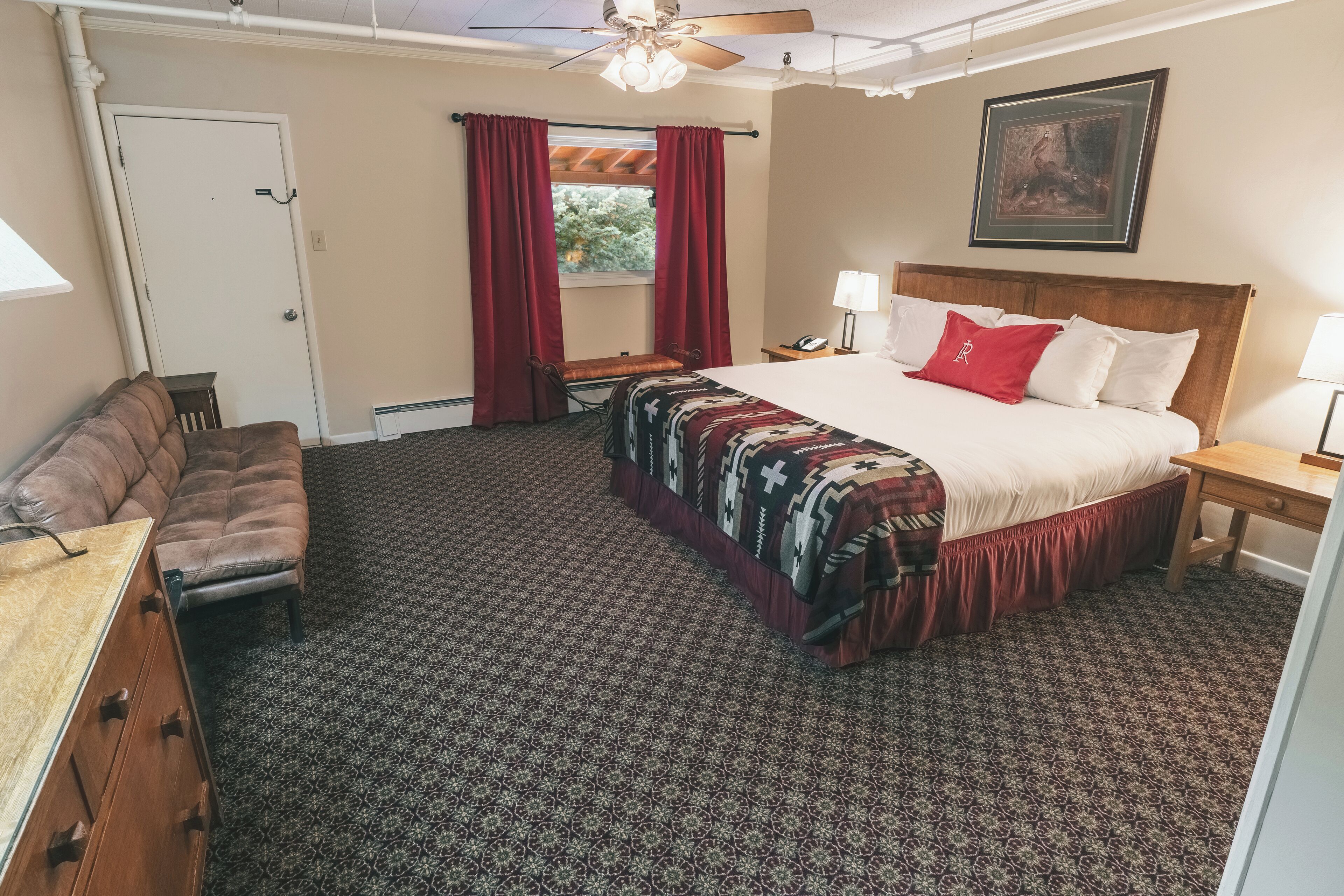 A hotel room with a bed, a desk, a chair, a window with red curtains, a ceiling fan, and a painting on the wall.