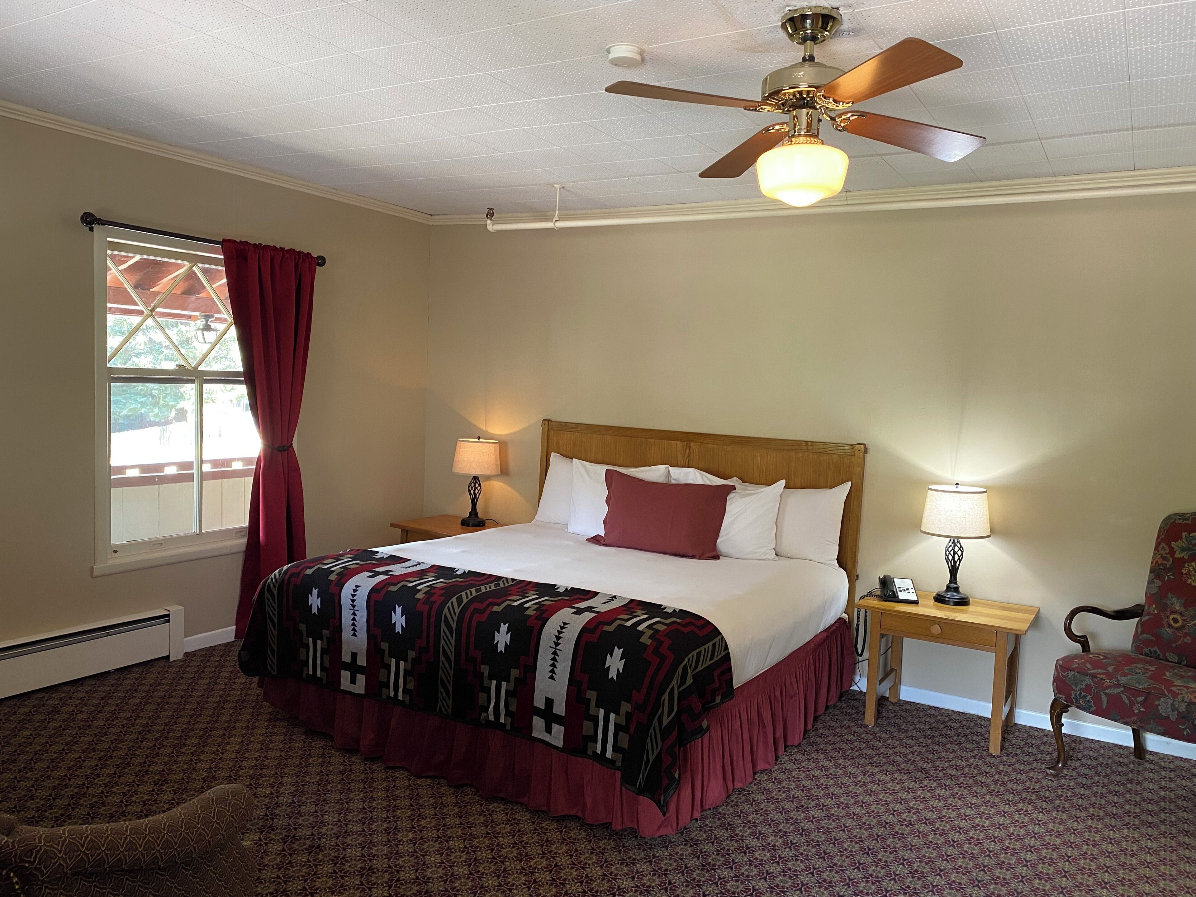 Habitación de hotel con una cama, dos mesitas de noche con lámparas, una silla, ventilador de techo y ventana con cortinas.