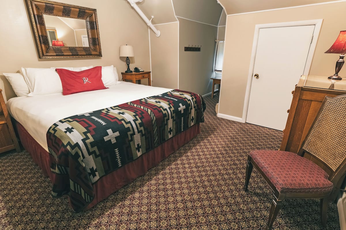 A hotel room with a large bed, a patterned carpet, a red chair, a wooden nightstand, a lamp, and a mirror on the wall.