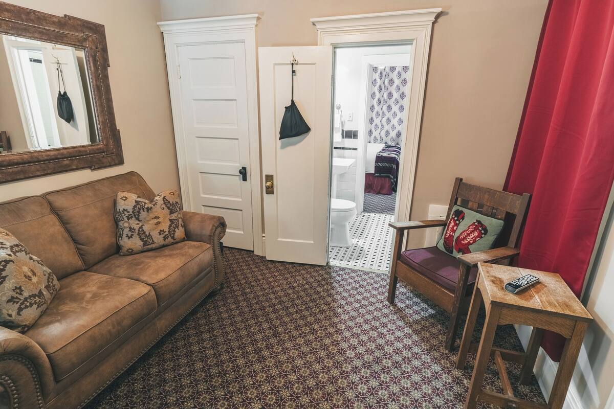 A living room with a brown sofa, patterned cushions, a wooden chair, a small table with a remote control, and a patterned carpet.