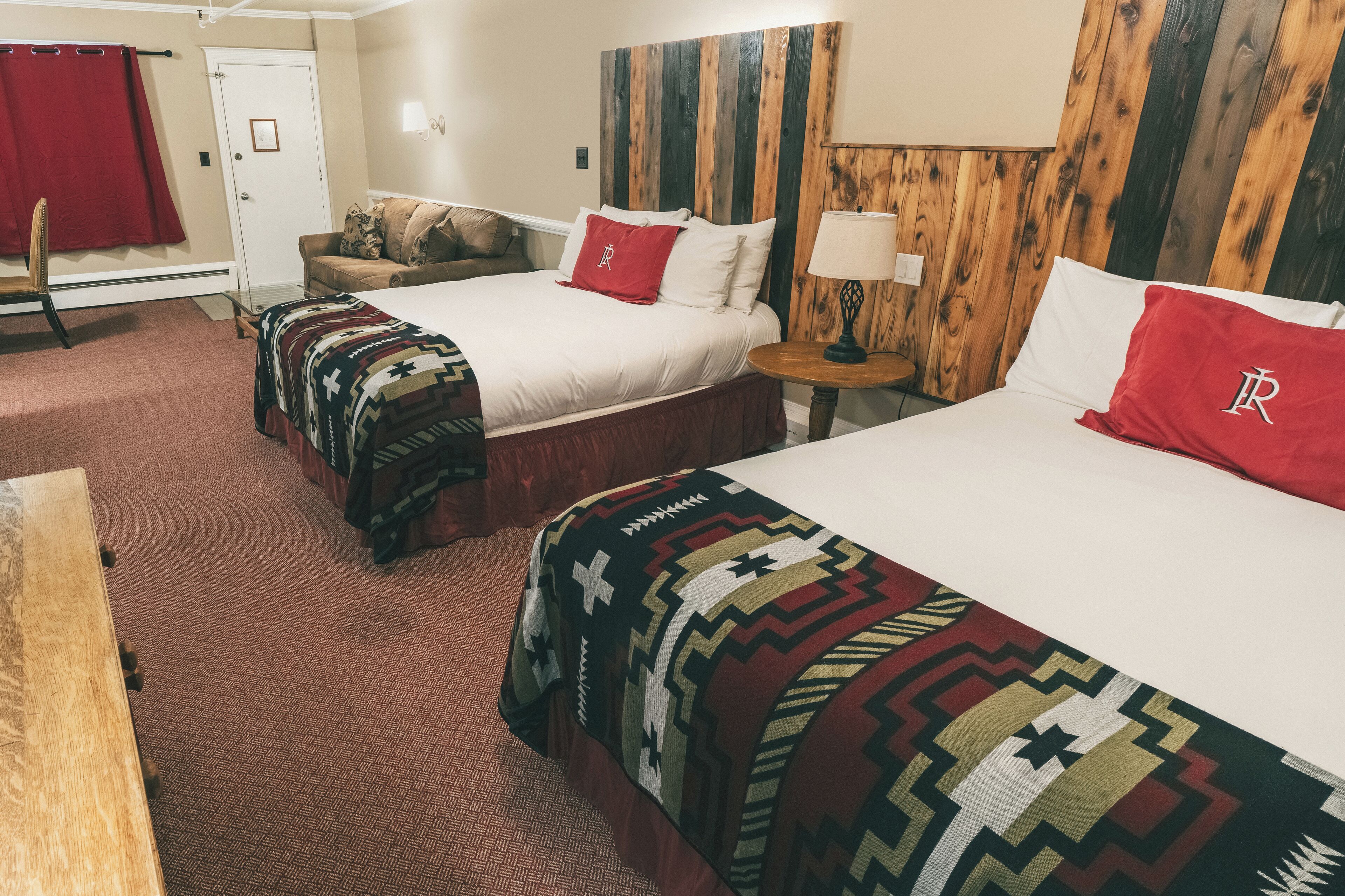 A hotel room with two beds, a wooden headboard, a red curtain, a small table with a lamp, and a patterned blanket.