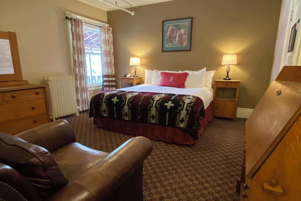 A hotel room with a bed, two bedside tables with lamps, a wooden dresser, a leather armchair, a radiator, a window with curtains, and a painting on the wall.