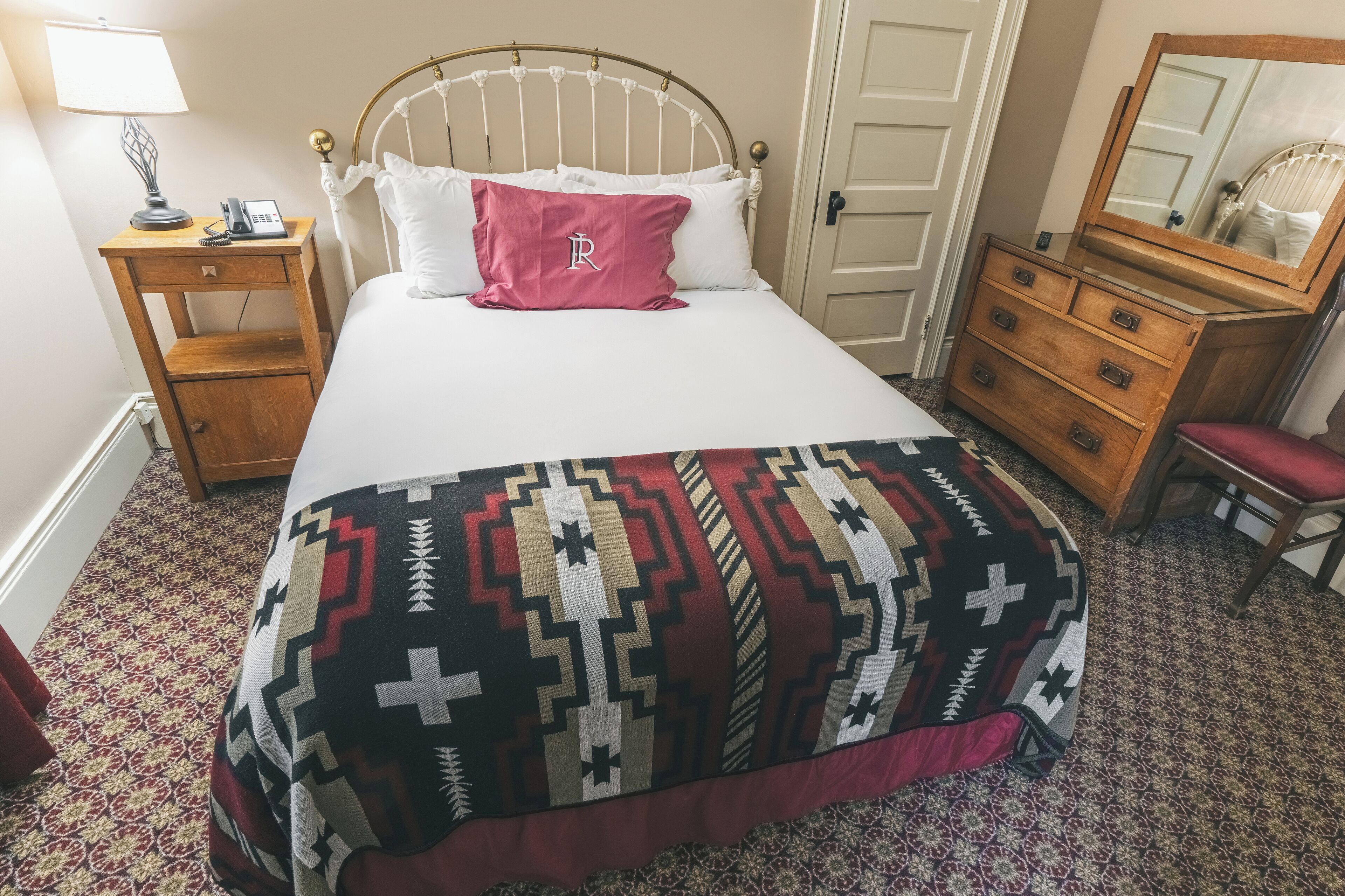 Een net opgemaakt bed met een geborduurde deken, een nachtkastje met lamp en telefoon, een houten dressoir met spiegel, en een deur op de achtergrond.