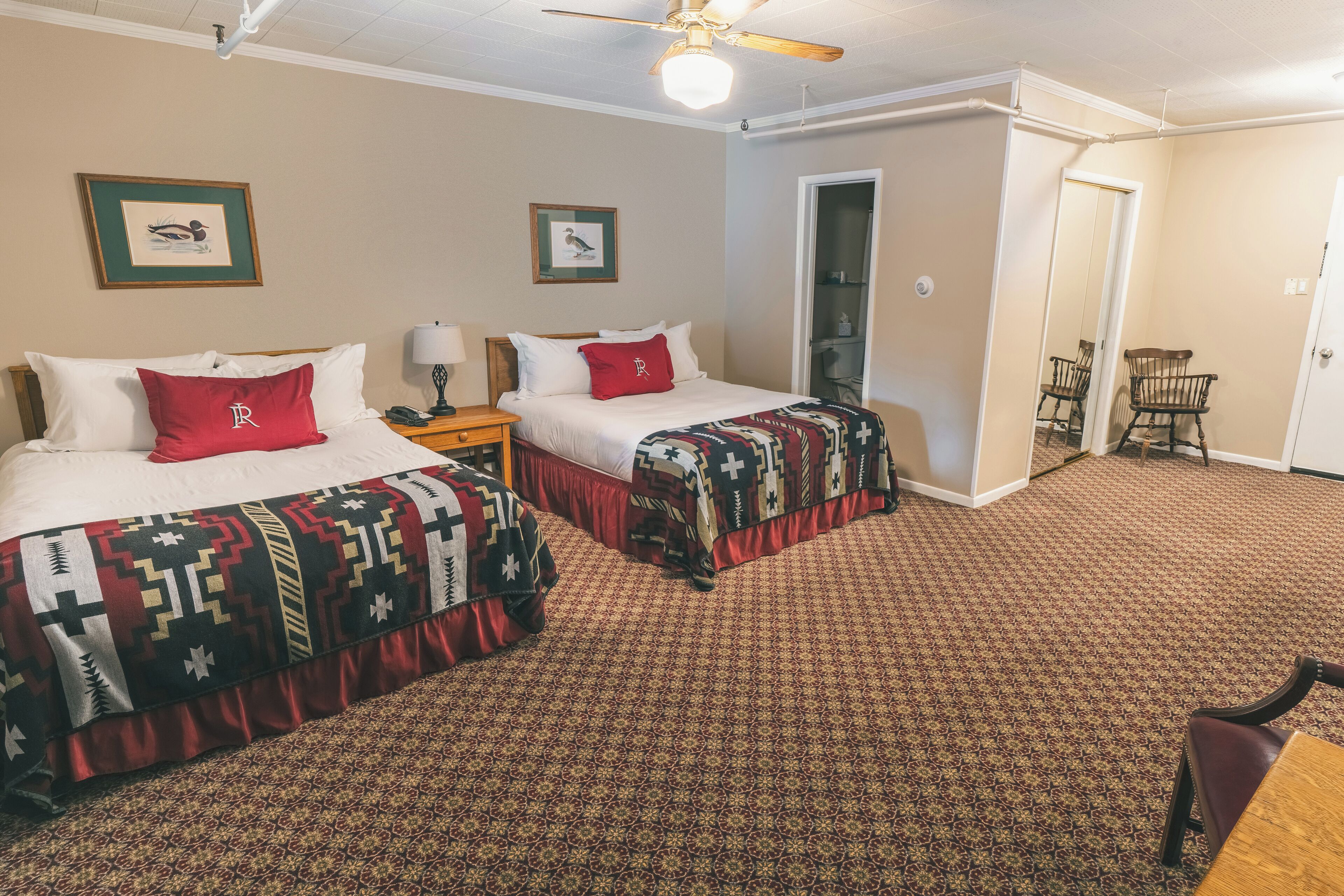 Een hotelkamer met twee bedden, elk met een bedovertrek met patroon en een rood kussen. Er is een nachtkastje met een lamp, een stoel en een deur naar een andere kamer.