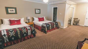 Een hotelkamer met twee bedden, elk met een bedovertrek met patroon en een rood kussen. Er is een nachtkastje met een lamp, een stoel en een deur naar een andere kamer.