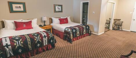 A hotel room with two beds, each with a patterned bedspread and a red pillow. There is a nightstand with a lamp, a chair, and a door leading to another room.