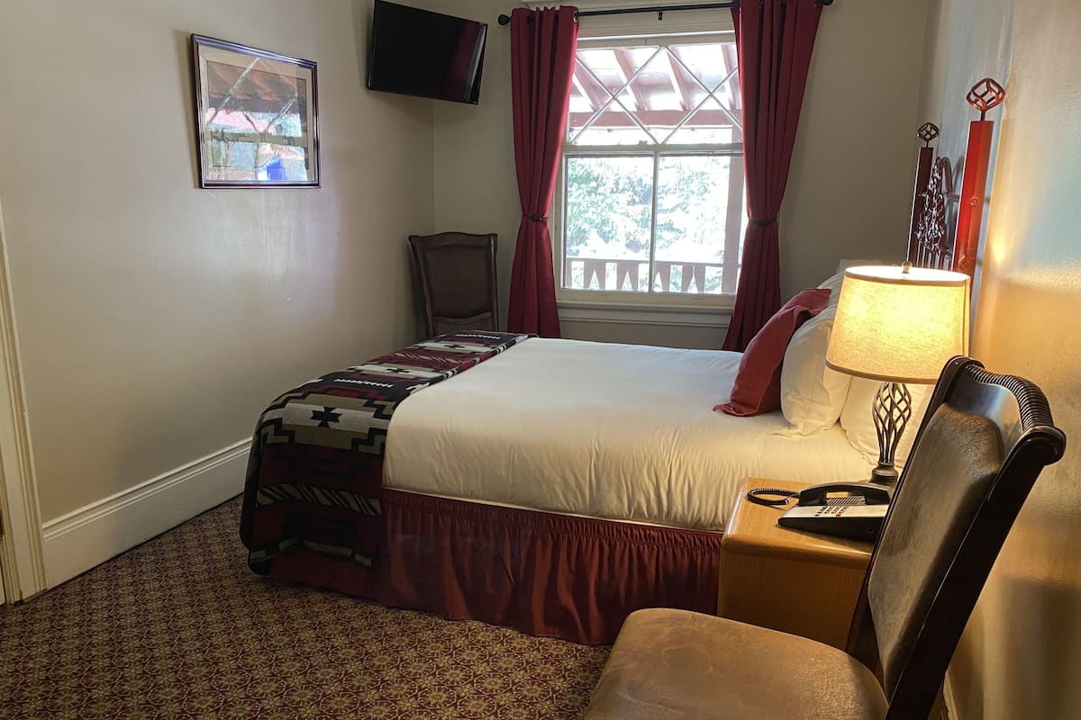 A hotel room with a bed, a chair, a desk with a lamp, a television on the wall, and a window with curtains.