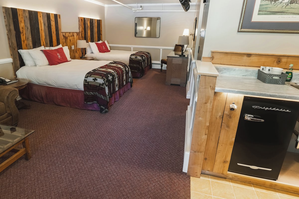 A hotel room with two beds, a sofa, a small table, a refrigerator, and a bathroom.