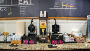 A waffle station with waffle batter dispensers and a waffle iron, along with a coffee machine and a stack of disposable cups. - Par-A-Dice Inn (East Peoria)