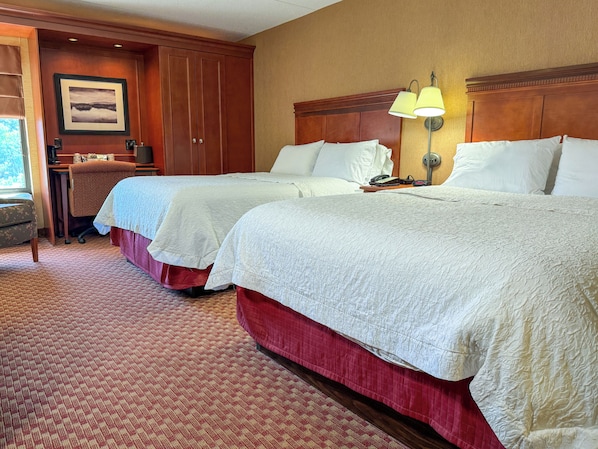 A hotel room with two beds, a desk, a chair, and a lamp.
