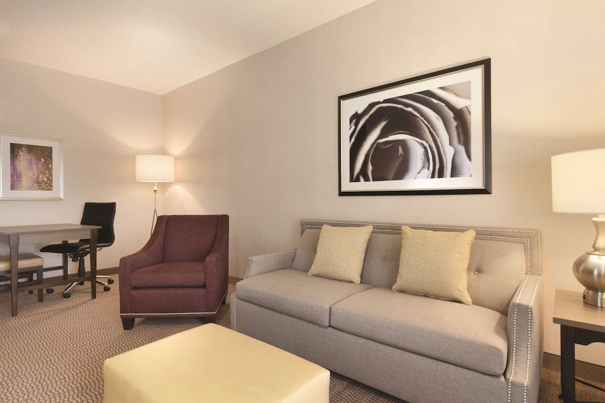 A hotel room with a grey sofa, a red armchair, a small table, a desk, and a framed abstract painting on the wall.