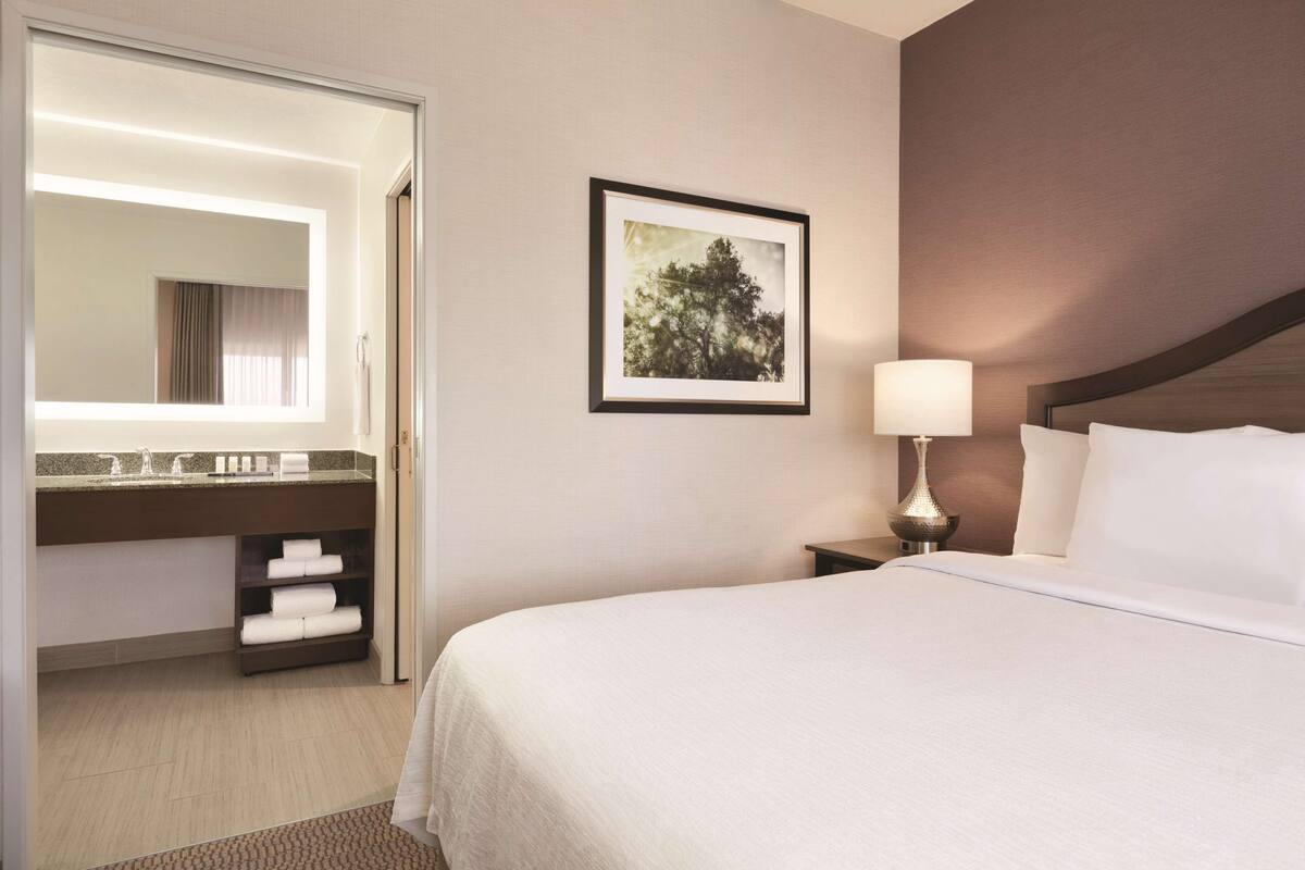 A hotel room with a large bed, a bedside table with a lamp, a framed picture on the wall, a bathroom with a mirror, and a towel rack.