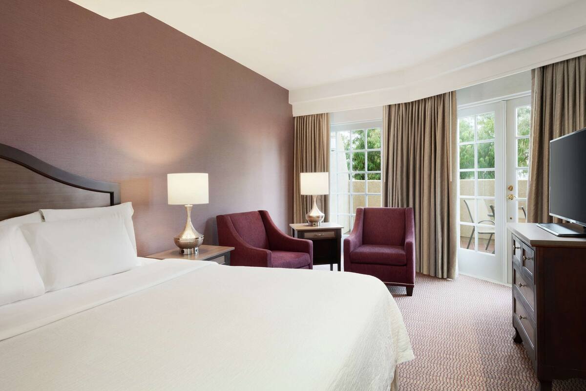 A hotel room with a large bed, two armchairs, a desk with a lamp, a television, and a view of the outdoors through a window with curtains.