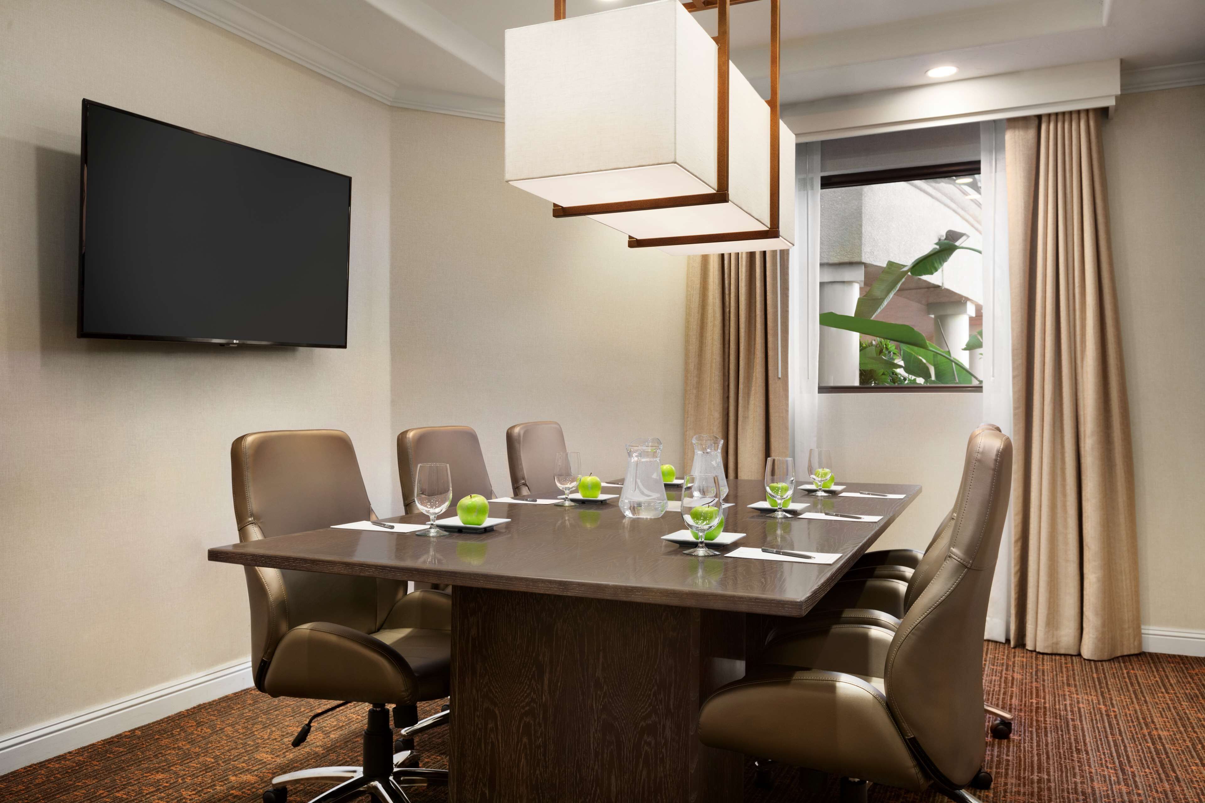 A conference room with a long table, six chairs, a flat-screen TV, and a window with curtains.