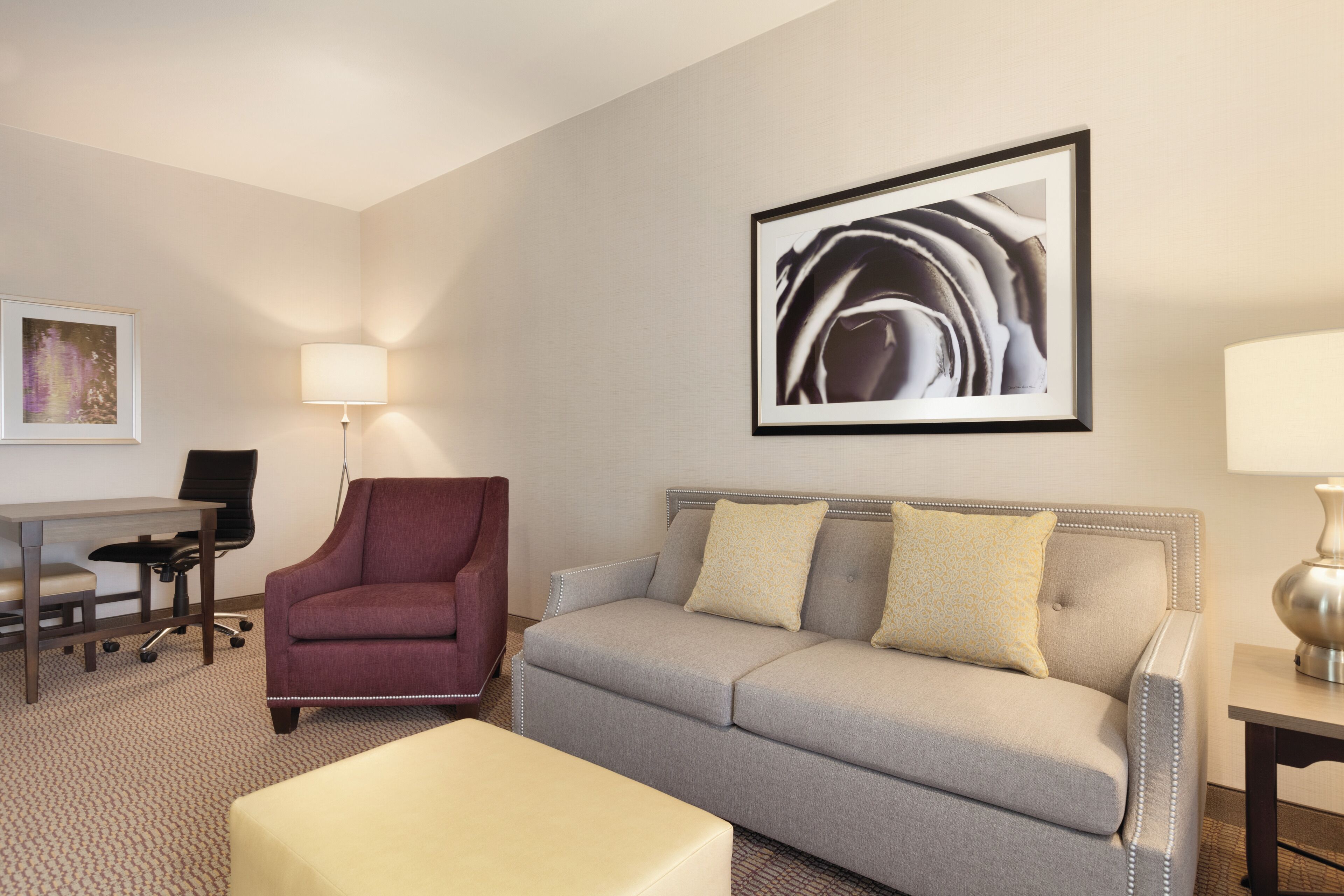 A hotel room with a grey sofa, a red armchair, a small table, a desk, and a chair.