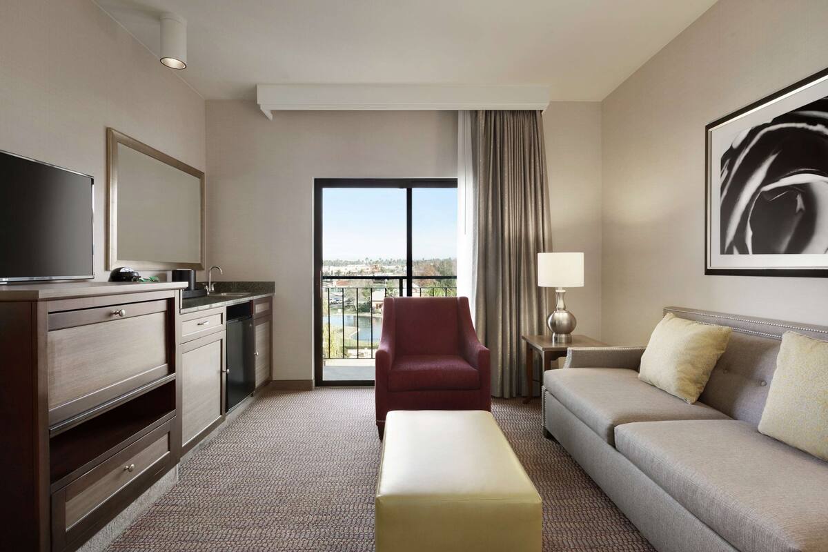 A hotel room with a sofa, a red armchair, a TV, a dining area, and a large window with a view.