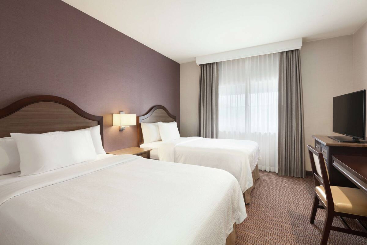 A hotel room with two beds, a desk, a chair, a television, and a window with curtains.