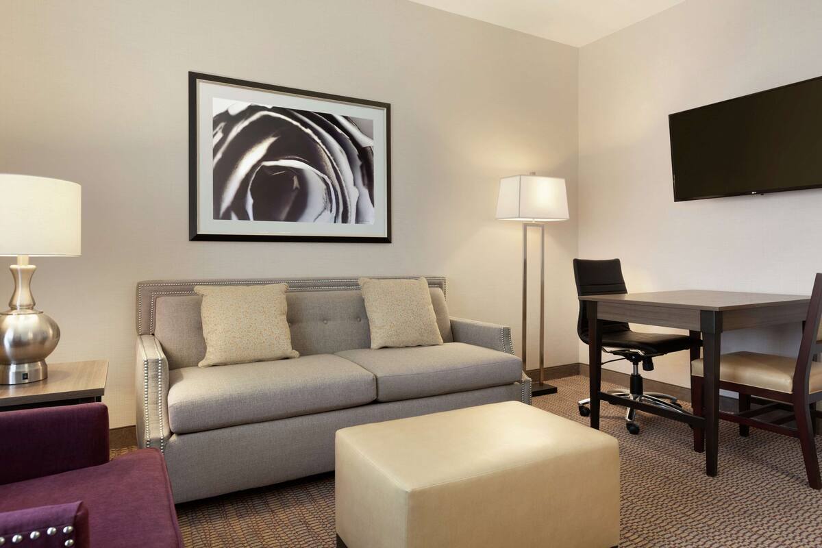 A hotel room with a grey sofa, a small table with a lamp, a desk with a chair, and a flat-screen TV on the wall.