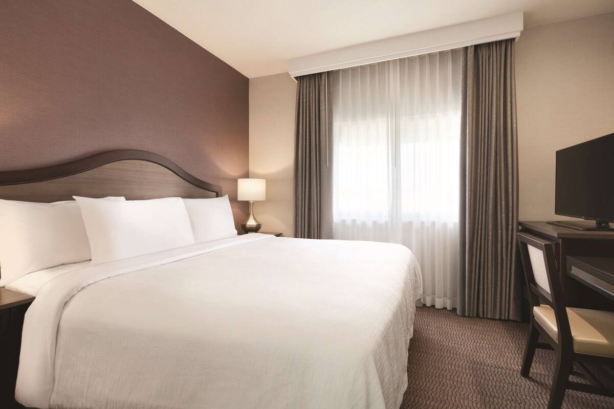 A hotel room with a large bed, a desk with a chair, a television, and a window with curtains.
