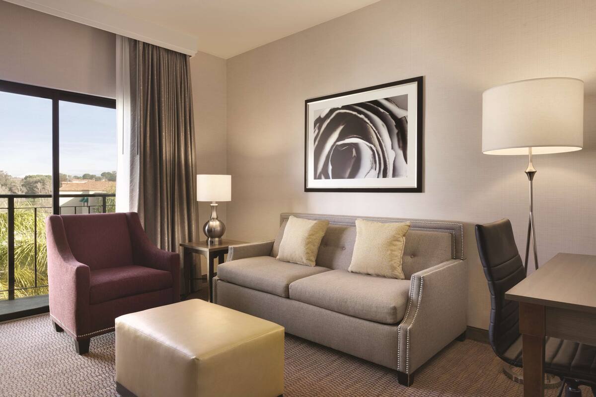 A hotel room with a large window, a sofa, a chair, a table, and a lamp.