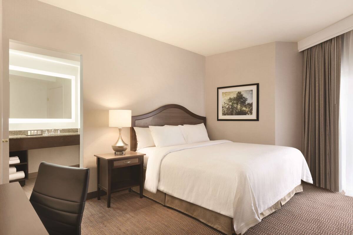 A hotel room with a bed, a desk with a chair, a nightstand with a lamp, a mirror, and a framed picture on the wall.