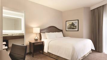 A hotel room with a bed, a desk with a chair, a nightstand with a lamp, a mirror, and a framed picture on the wall.