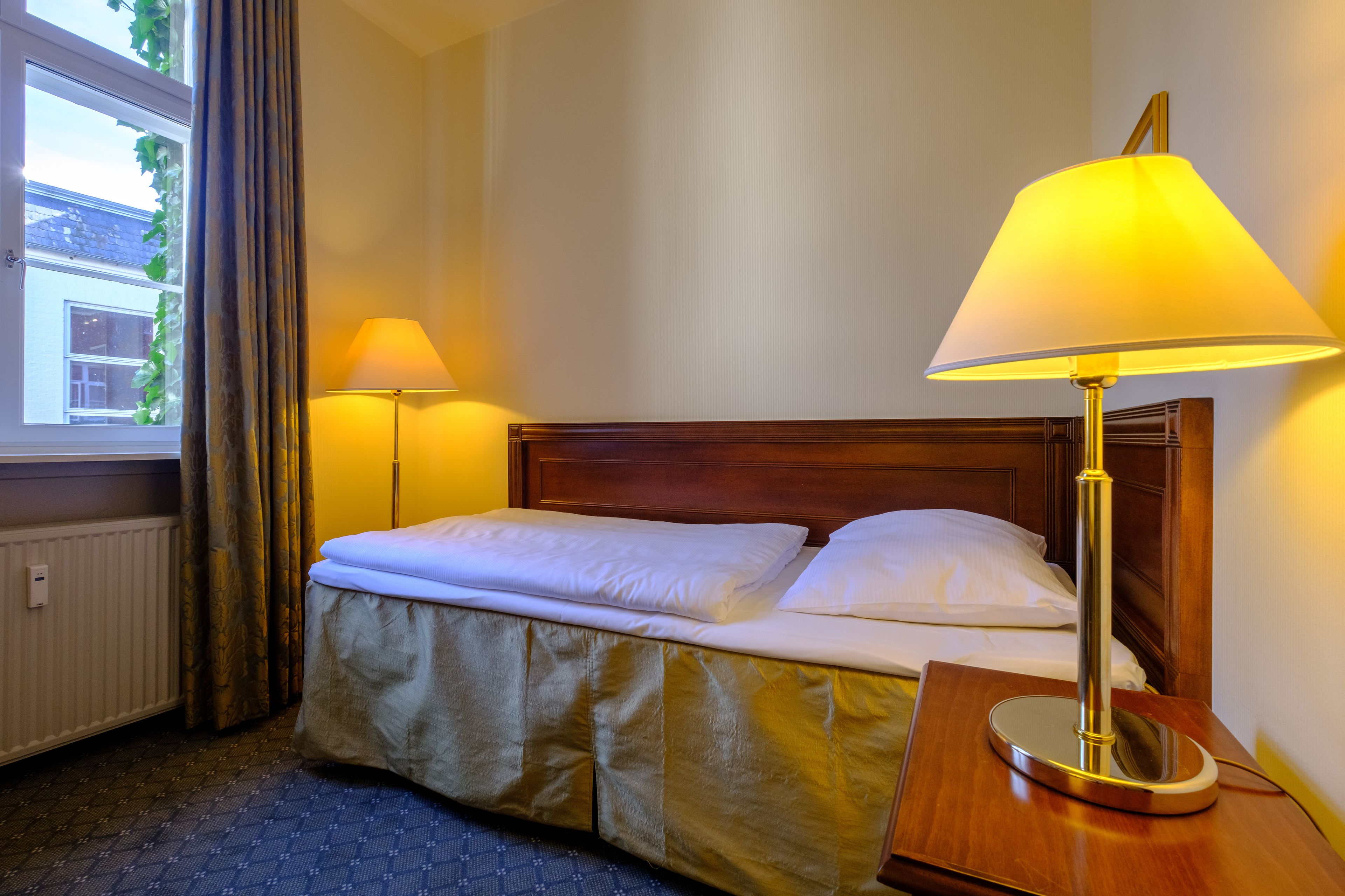 Una habitación de hotel con una cama, dos lámparas de noche, un cabecero de madera y una ventana con cortinas.
