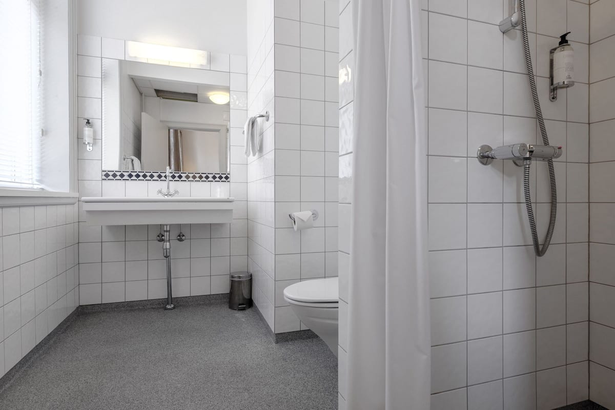 Un baño con paredes de azulejos blancos, un lavamanos con espejo, un inodoro y una ducha con manguera.