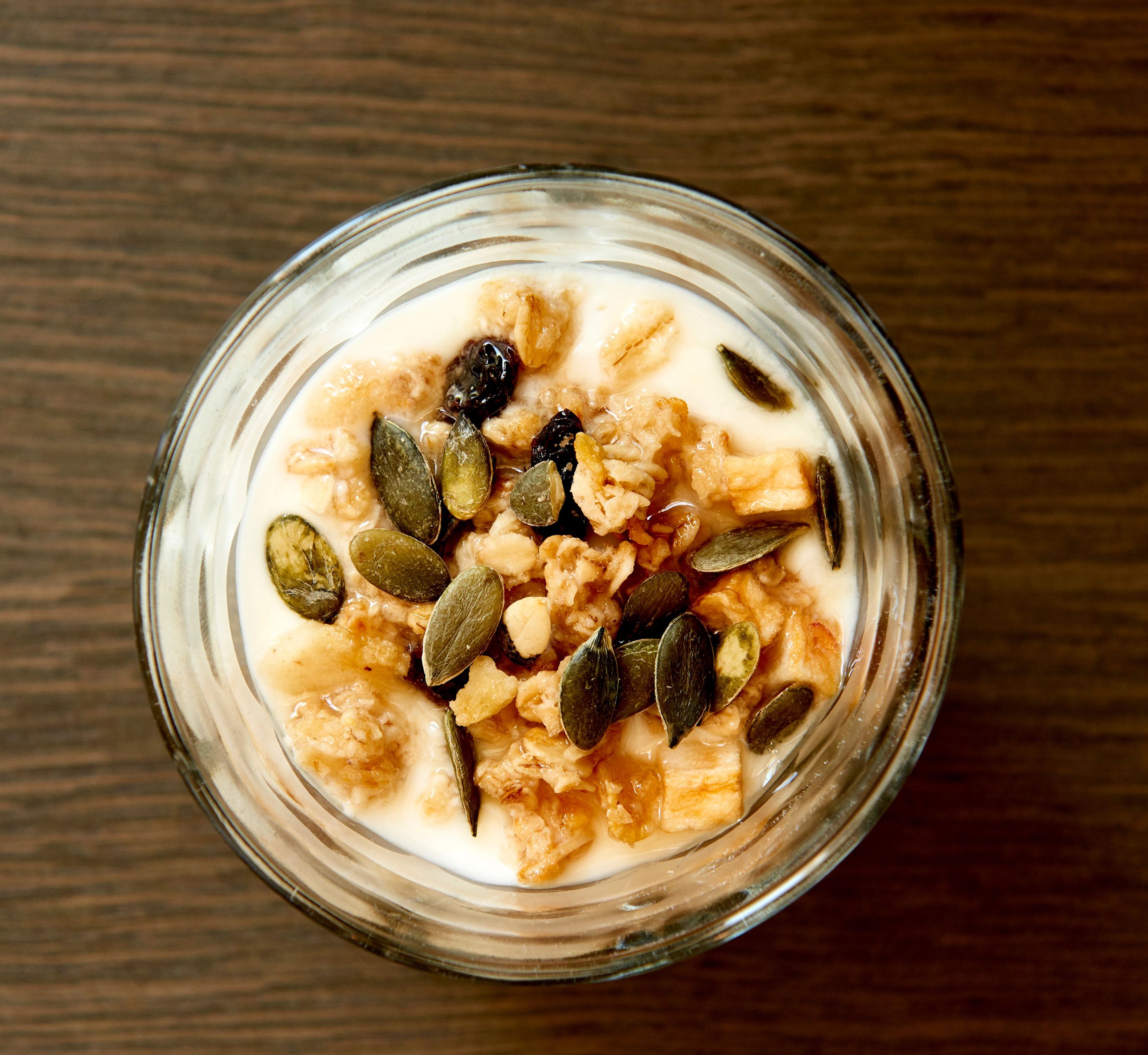 A glass bowl of oatmeal with pumpkin seeds and raisins on a wooden surface.