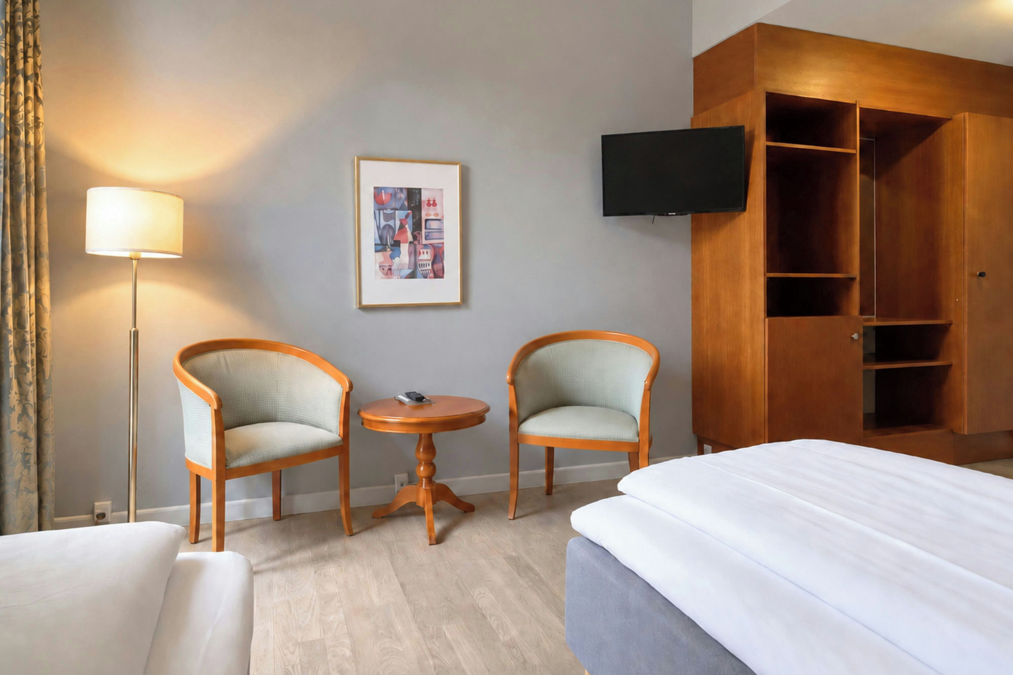 A hotel room with a bed, two armchairs, a small table, a lamp, a TV, and a wardrobe.