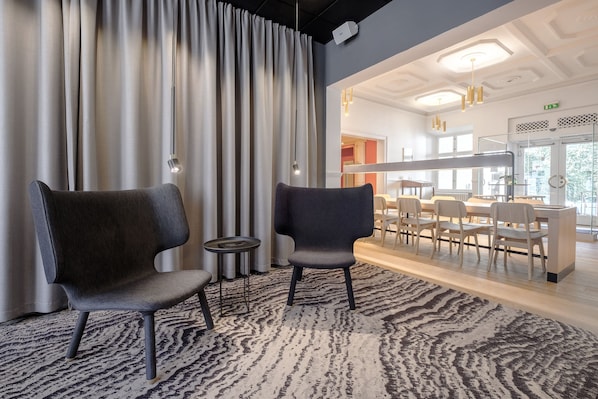 Un moderno vestíbulo de hotel con dos sillas oscuras, una alfombra estampada y vistas a un comedor con sillas de colores claros y una barra.