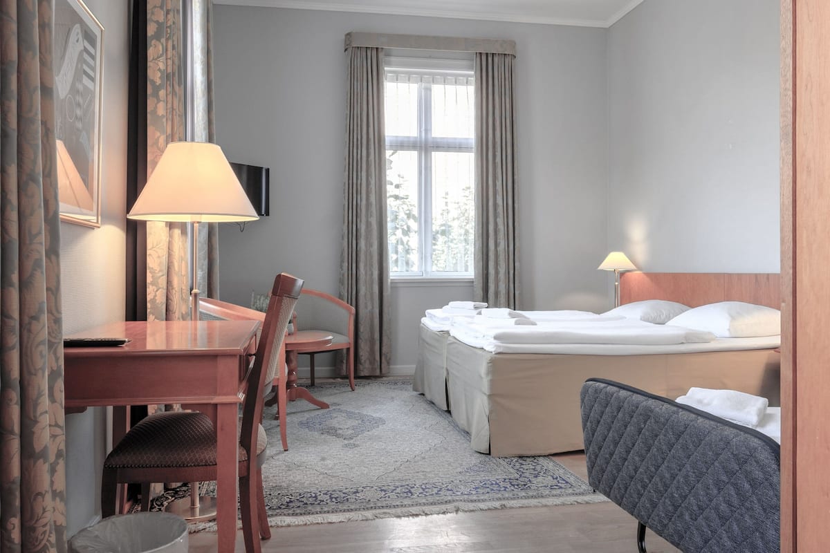 Habitación de hotel con cama, escritorio, silla, televisor y ventana con cortinas.