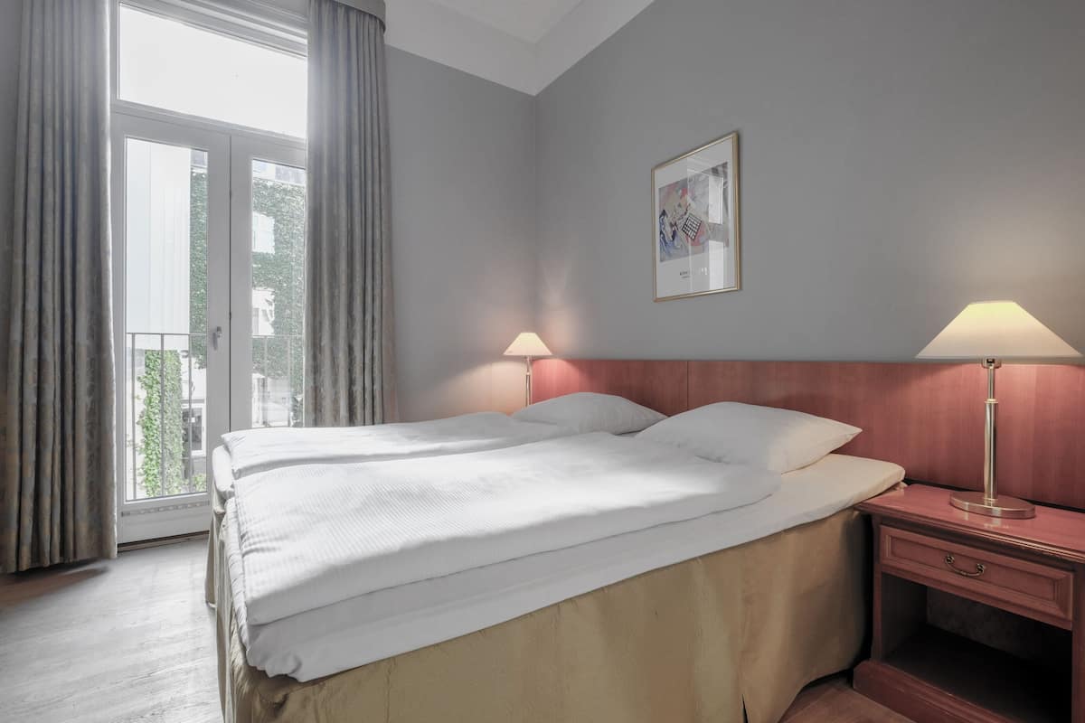Habitación de hotel con una cama grande, dos mesitas de noche, una ventana con cortinas y un cuadro en la pared.